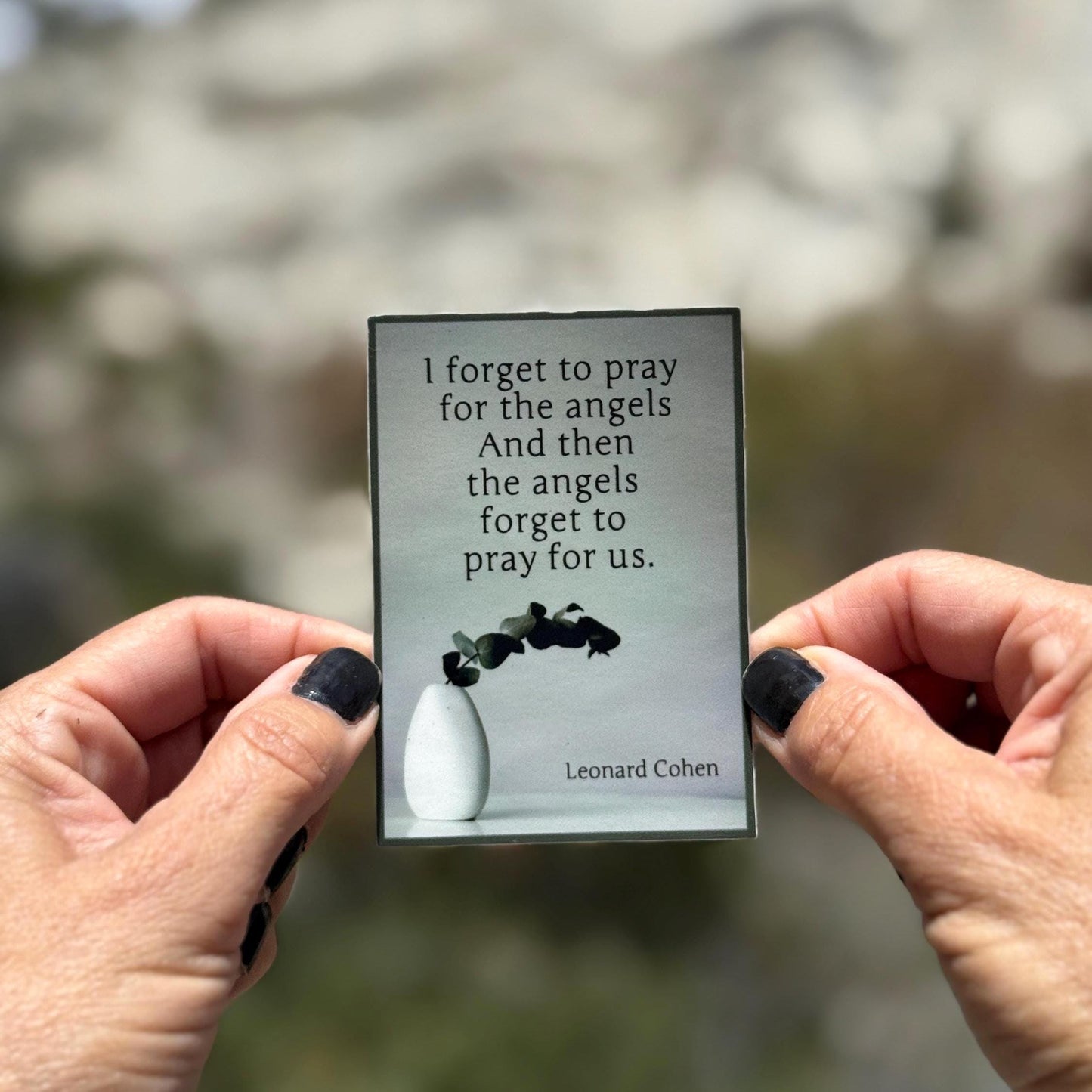 Holding a sticker with a quote by Leonard Cohen and a minimalistic illustration.
