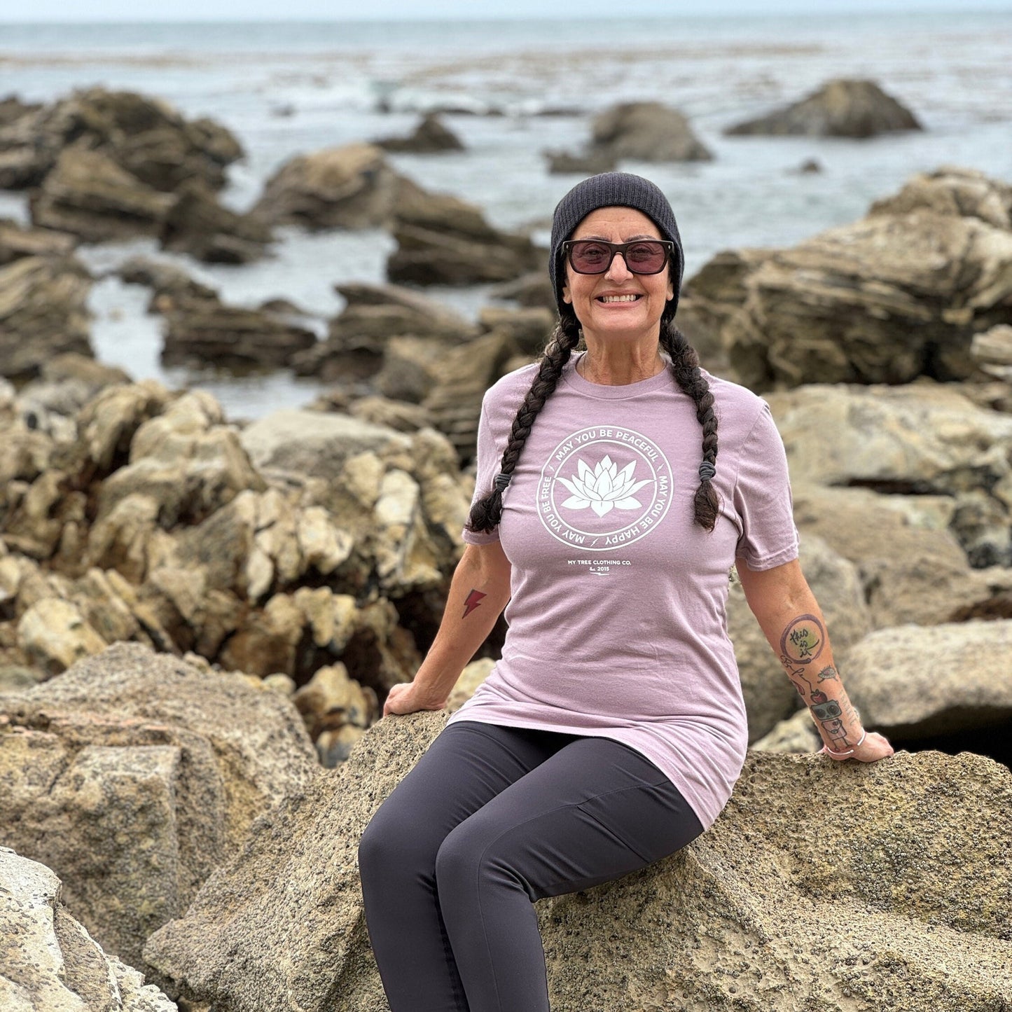 Person sitting on rocks by the ocean wearing a light purple t-shirt with a white lotus in the center and the words, may you be peaceful may you be happy may you be free.