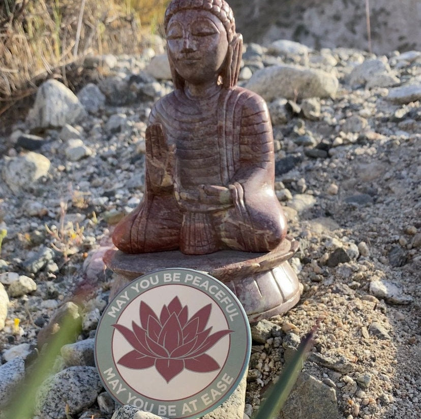 Buddha statue with a 3.5" lotus flower magnet with the metta prayer, 'may you be peaceful may you be at ease' on a rocky ground in the California desert.