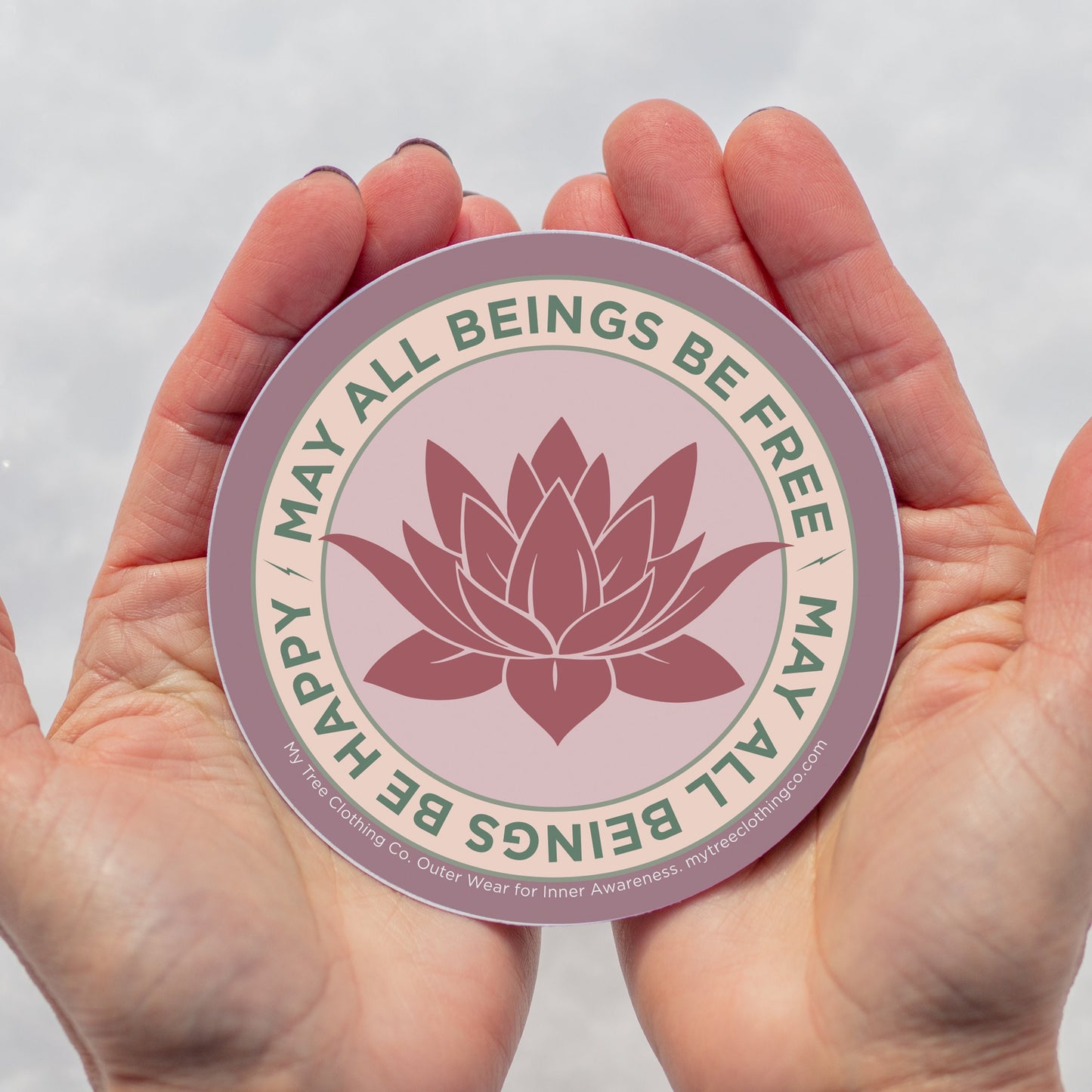4" Circular sticker with lotus flower design and the text, MAY ALL BEINGS BE FREE MAY ALL BEINGS BE HAPPY held between two hands against a white background.