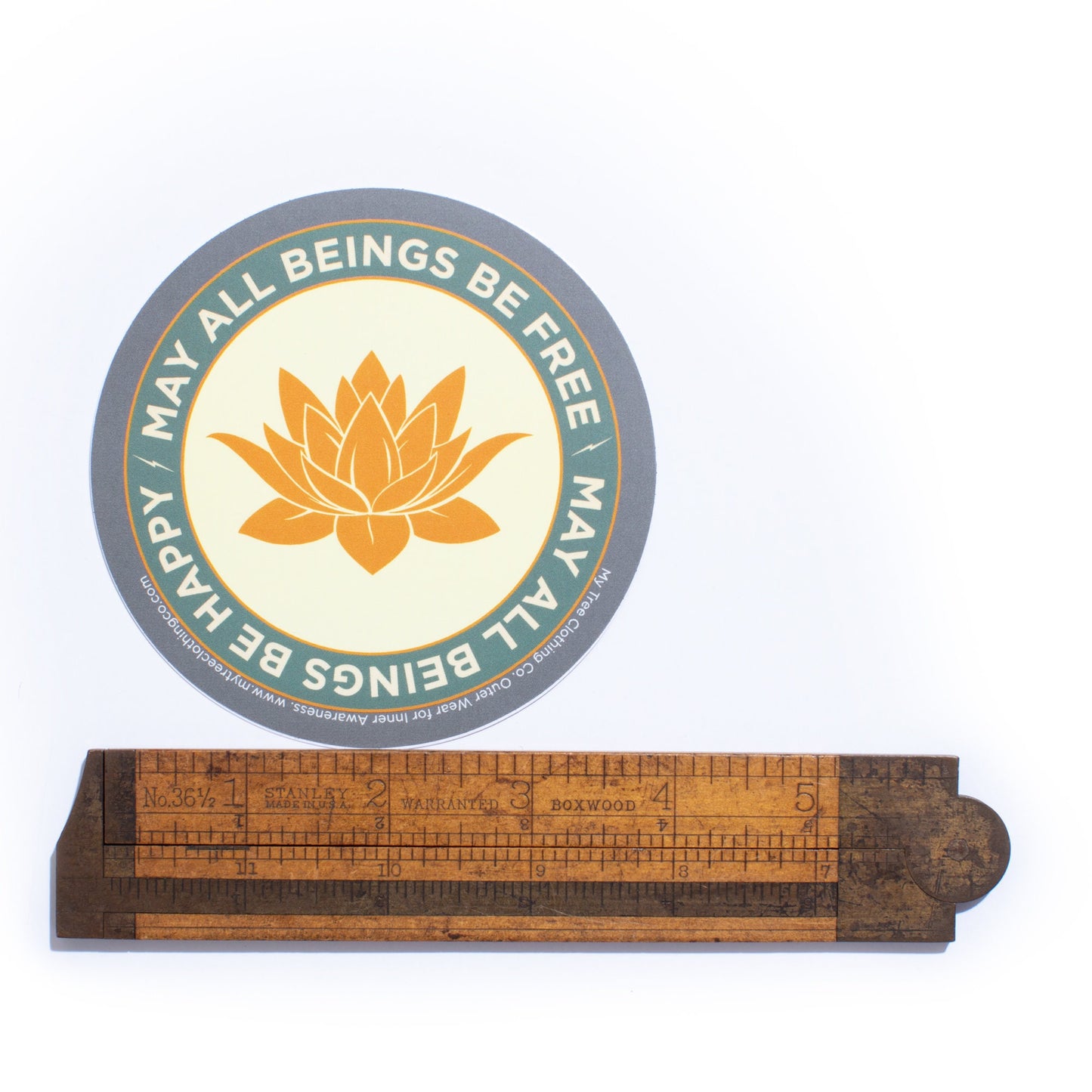 4" circular sticker with a saffron colored lotus flower design and Mettā prayer being measured by an antique wooden ruler.