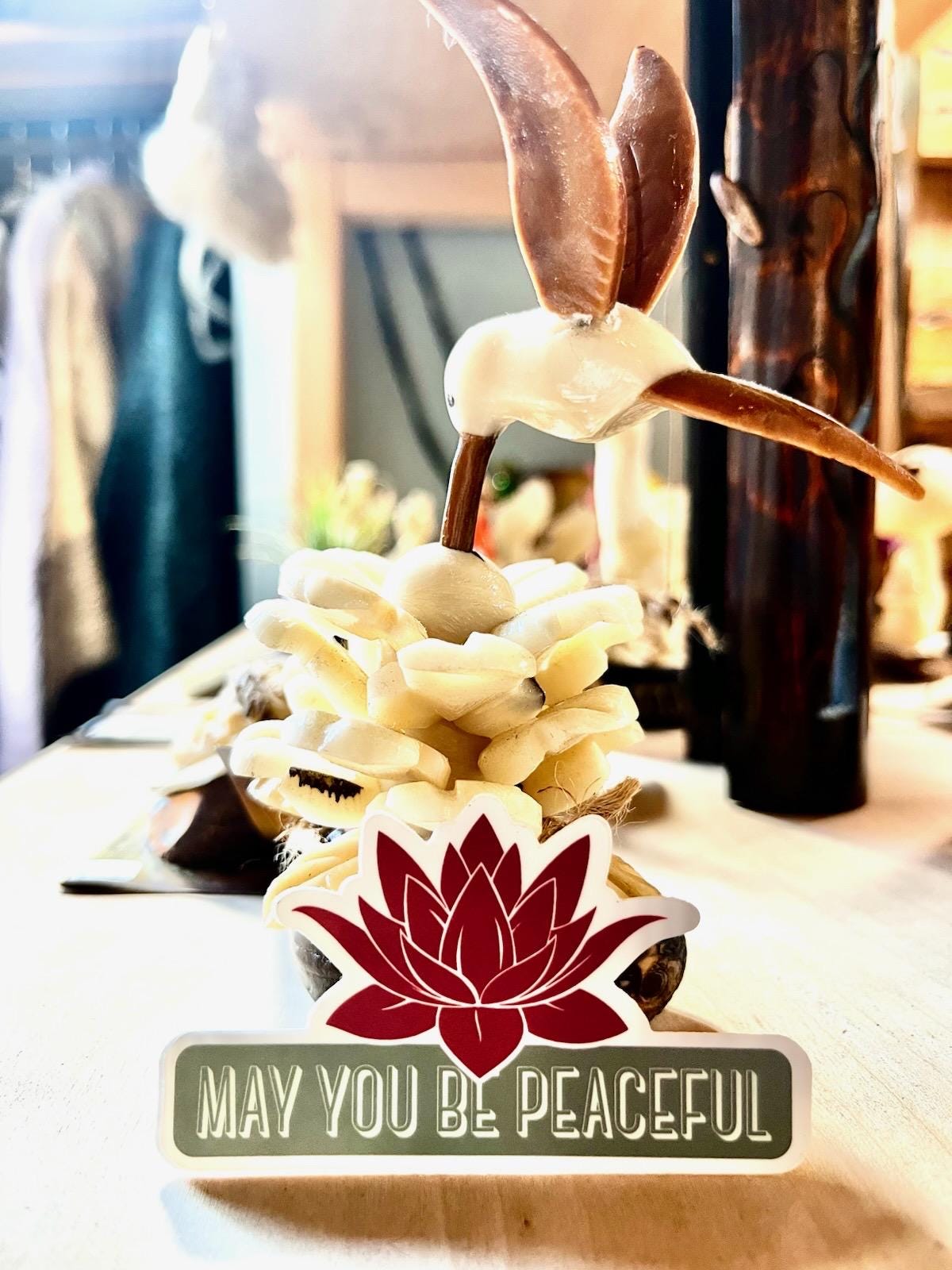 Decorative item with a lotus flower and text 'May you be peaceful' on a blurred background.