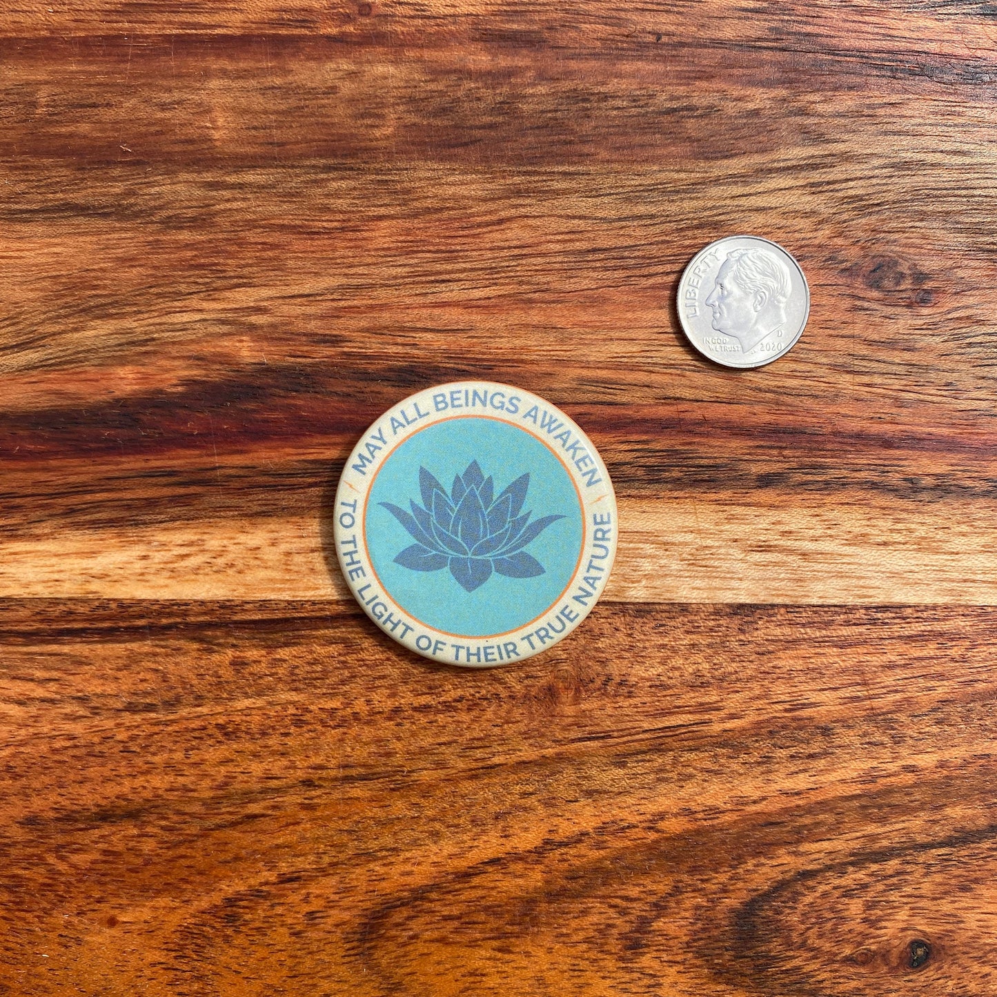 1.5" button with the mantra that reads, may all beings awaken to the light of their true nature, with a lotus design on wooden surface with coin for scale.