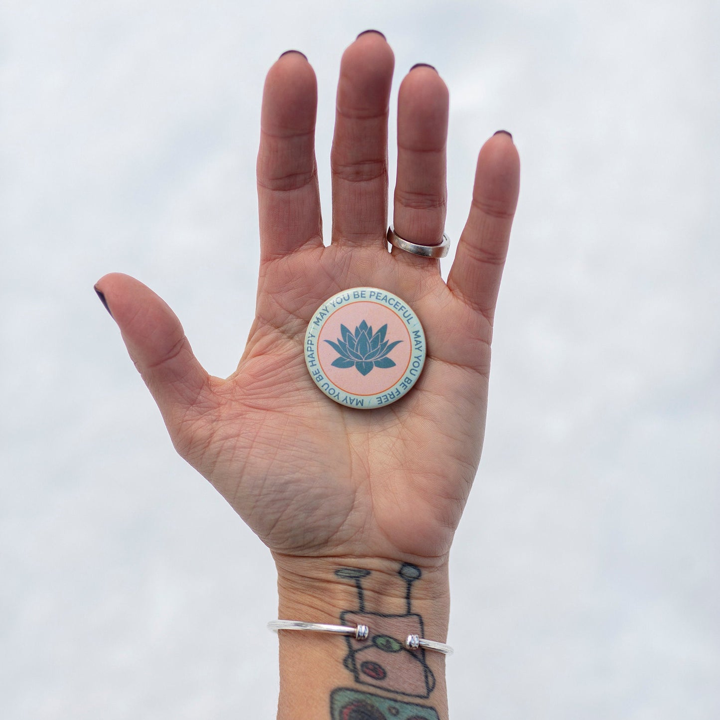 Hand holding a circular 1.5" button with a lotus flower design and the text, may you be happy may you be peaceful, may you be free, against a white background.