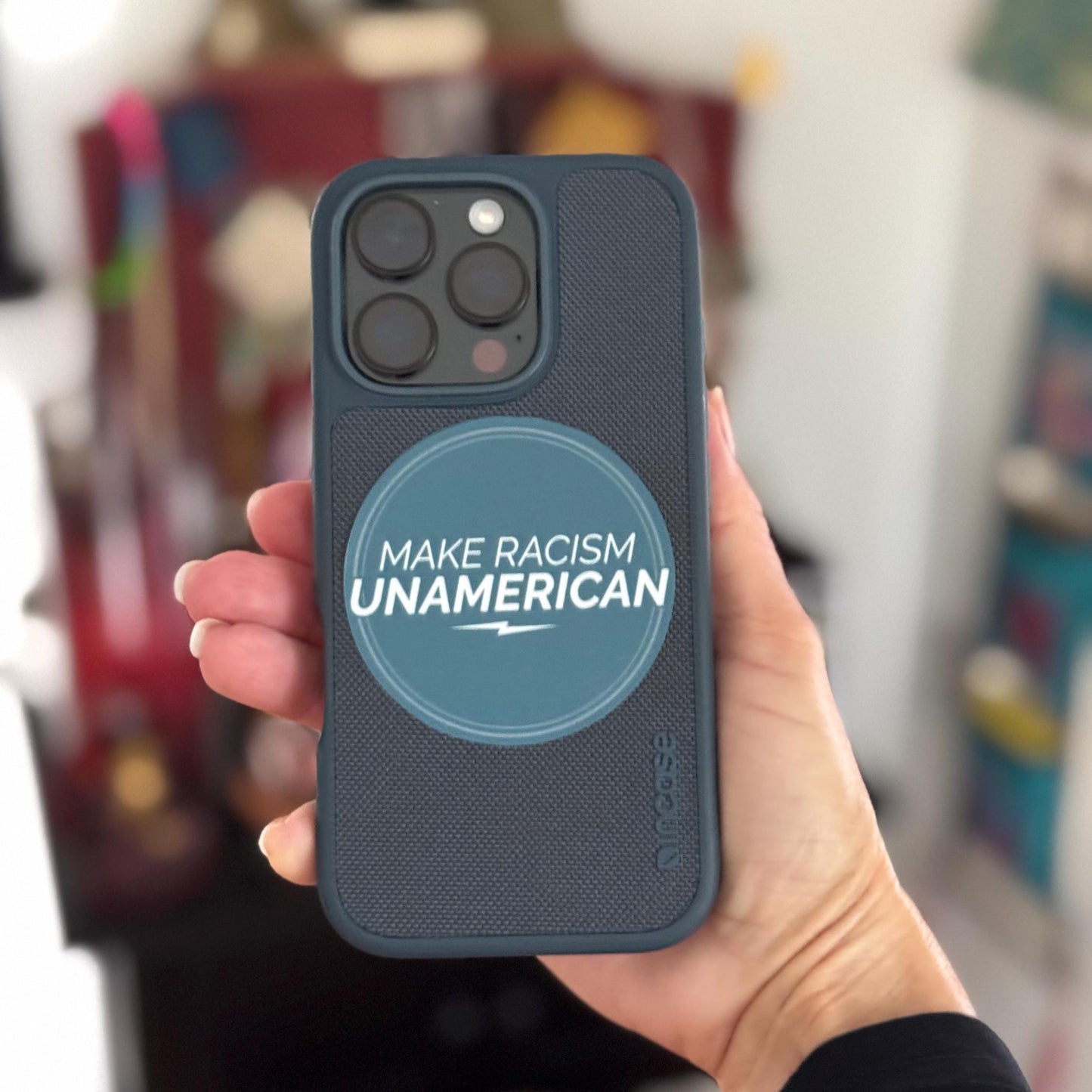 Phone case with 'Make Racism Unamerican' text held by a hand against a blurred background.
