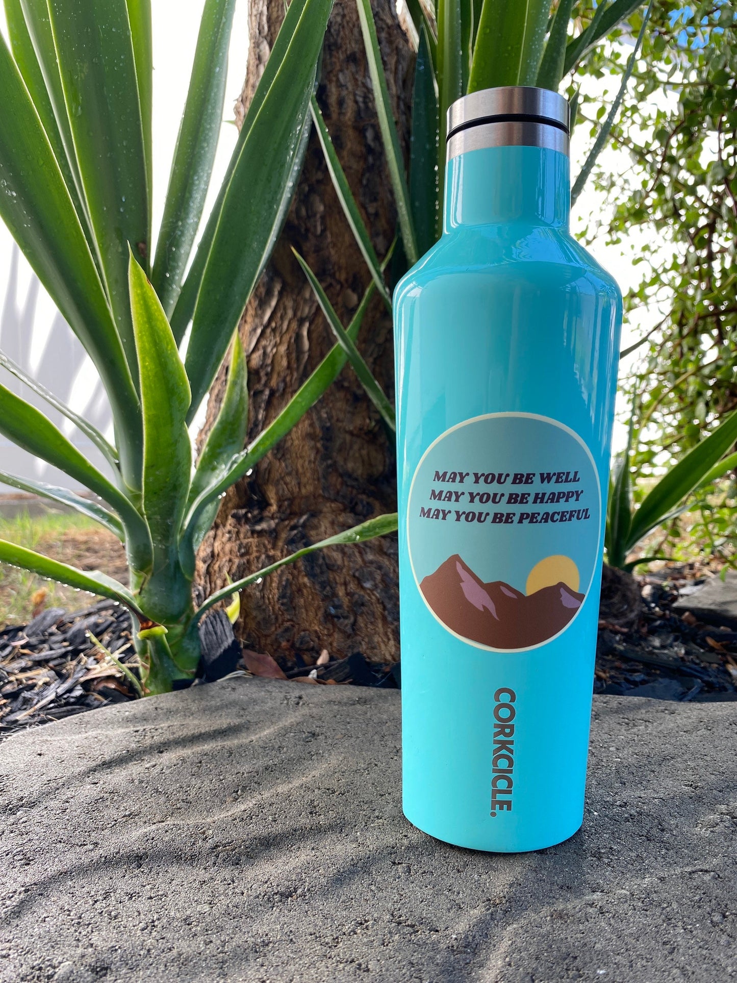 Blue Corkcicle water bottle with a Buddhist loving kindness mantra sticker that reads, may you be well may you be happy may you be peaceful, in a natural background.