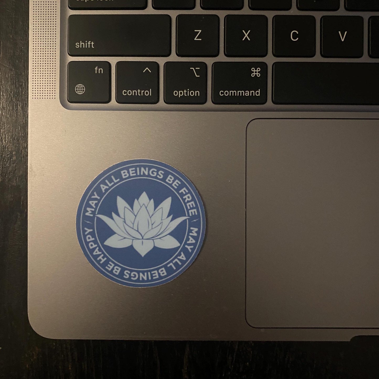 Laptop with a Buddhist meditation sticker in a gorgeous blue with a lotus flower design and the text, 'may all beings be free may all beings be happy' on the palm rest area.