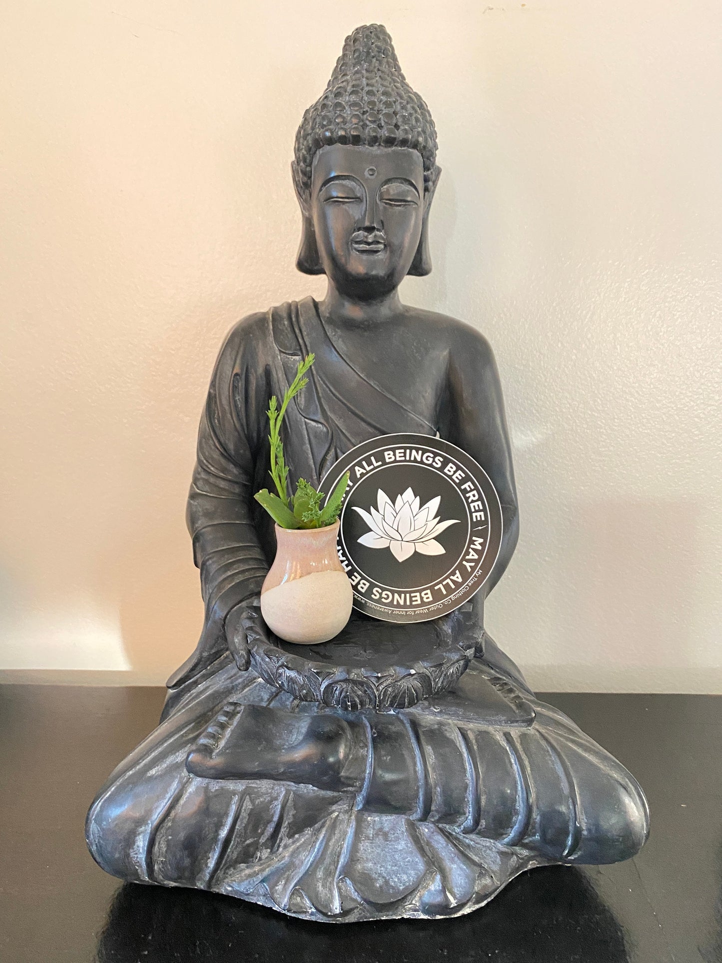 Buddha statue holding a pot with a plant and a 4" black and white circular sticker with a lotus flower design and text, MAY ALL BEINGS BE FREE MAY ALL BEINGS BE HAPPY