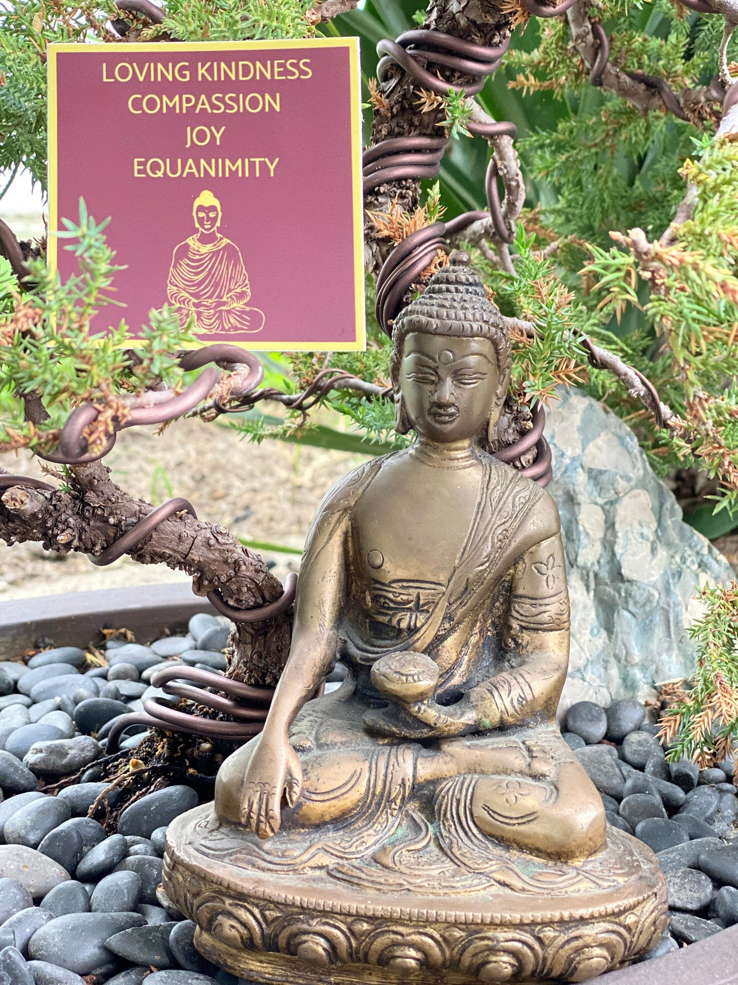 Bronze Buddha statue in a garden setting with a Tibetan red and gold three inch sticker with 'Loving Kindness, Compassion, Joy, Equanimity' text and a Buddha illustration.