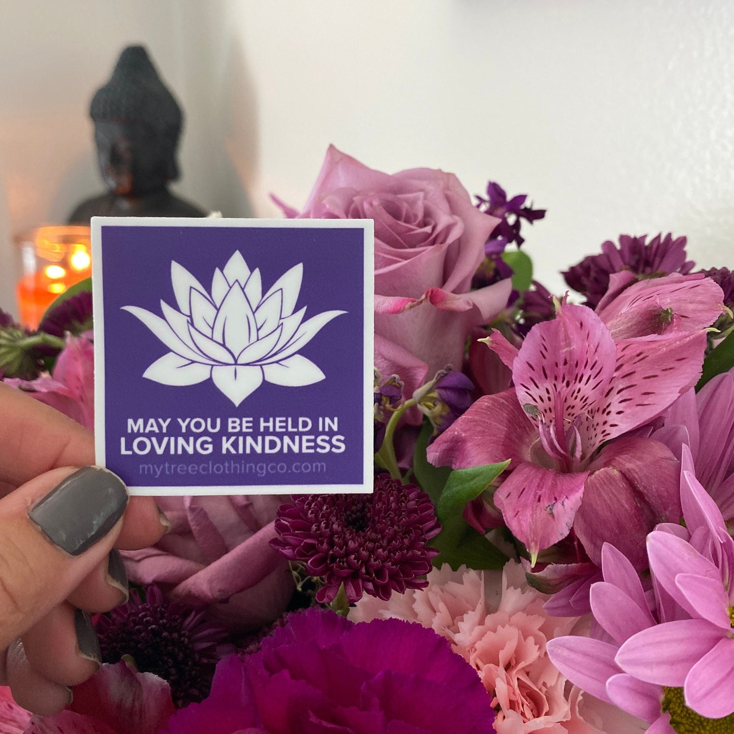 Person holding a 2" square purple and white lotus sticker with the words, may you be held in loving kindness, amidst flowers.