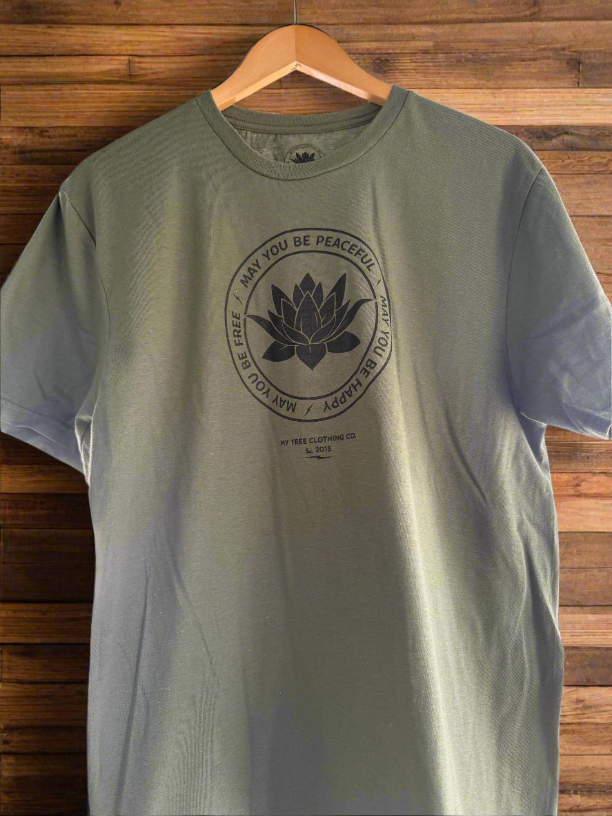 Green t-shirt with a black lotus flower design with the text, may you be peaceful, may you be happy, may you be free, on a wooden hanger against a wooden background.