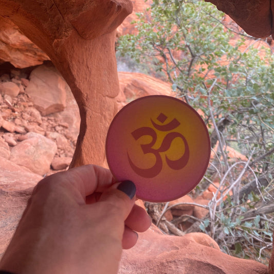 Hand holding a pink and gold 3" circular Om symbol sticker in front of red rocks and greenery in Nevada.