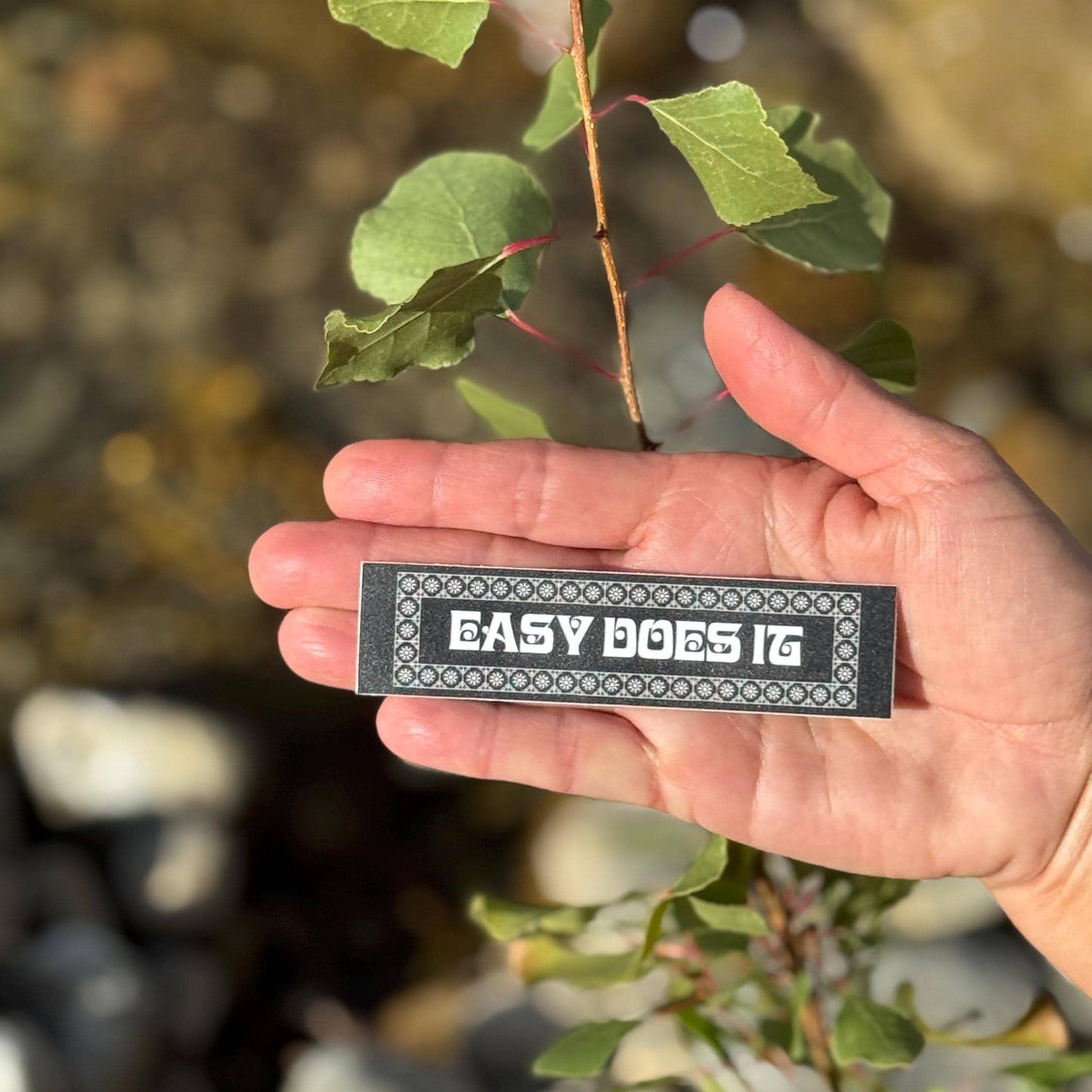 Hand holding a rectangular black and white 'Easy Does It' sticker with a natural background.