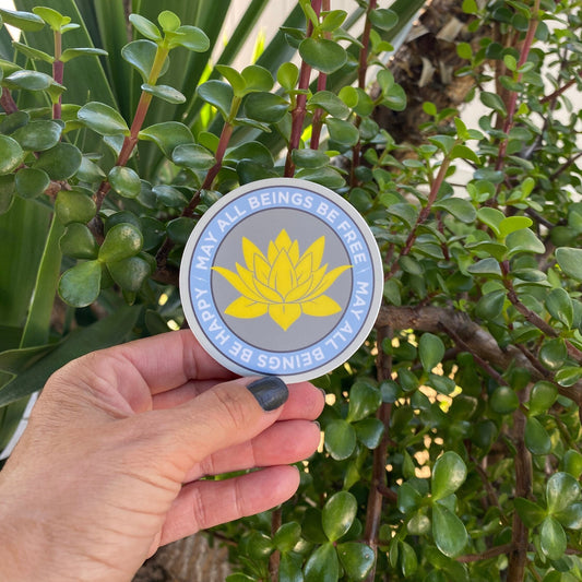 Hand holding a 3" circular sticker with a lotus flower design and text, may all beings be free may all beings be happy against a green leafy background.
