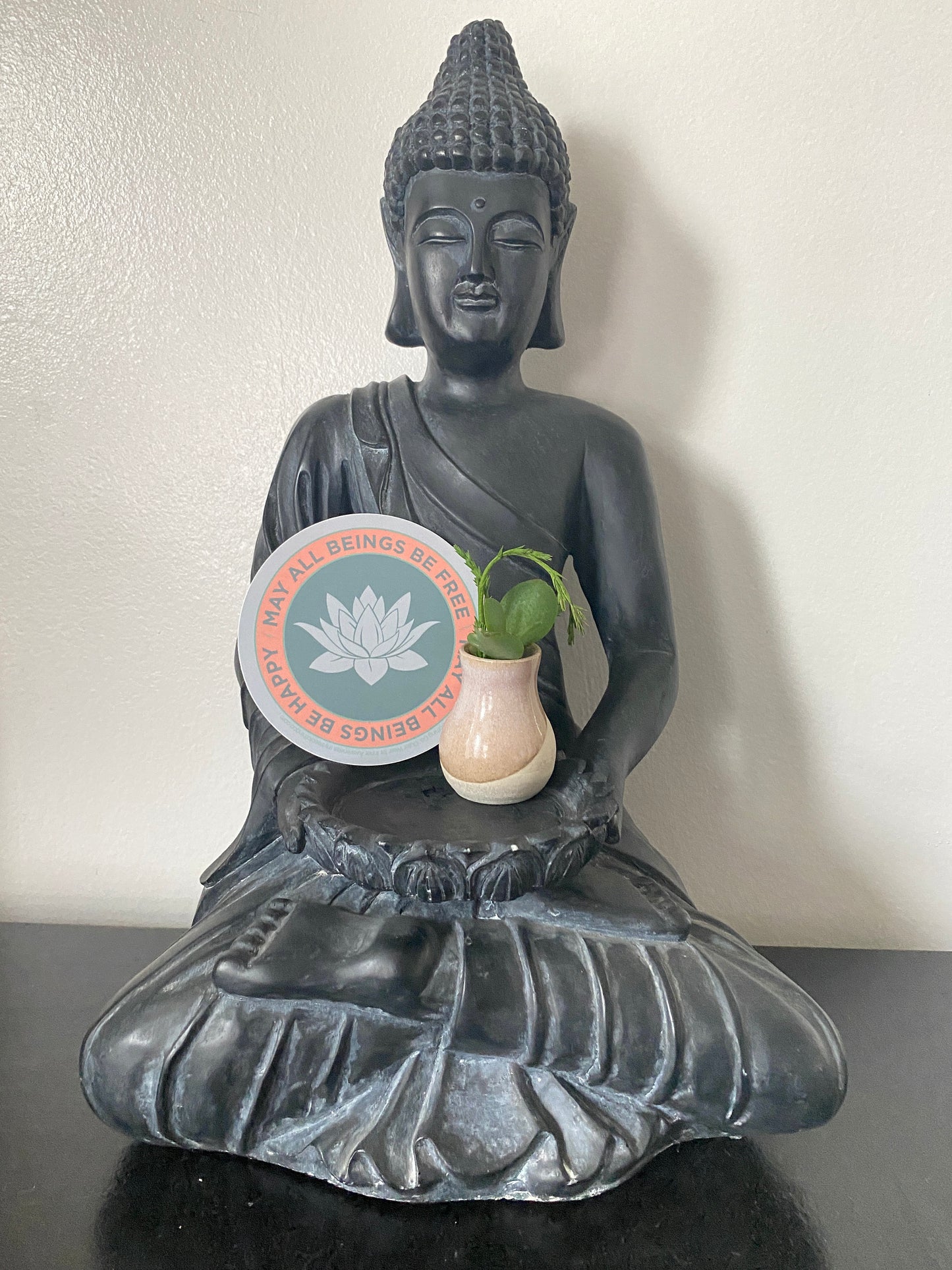 Statue of a Buddha holding a small plant and a 4" circular sticker with a lotus flower design and text, may all beings be free may all beings be happy.