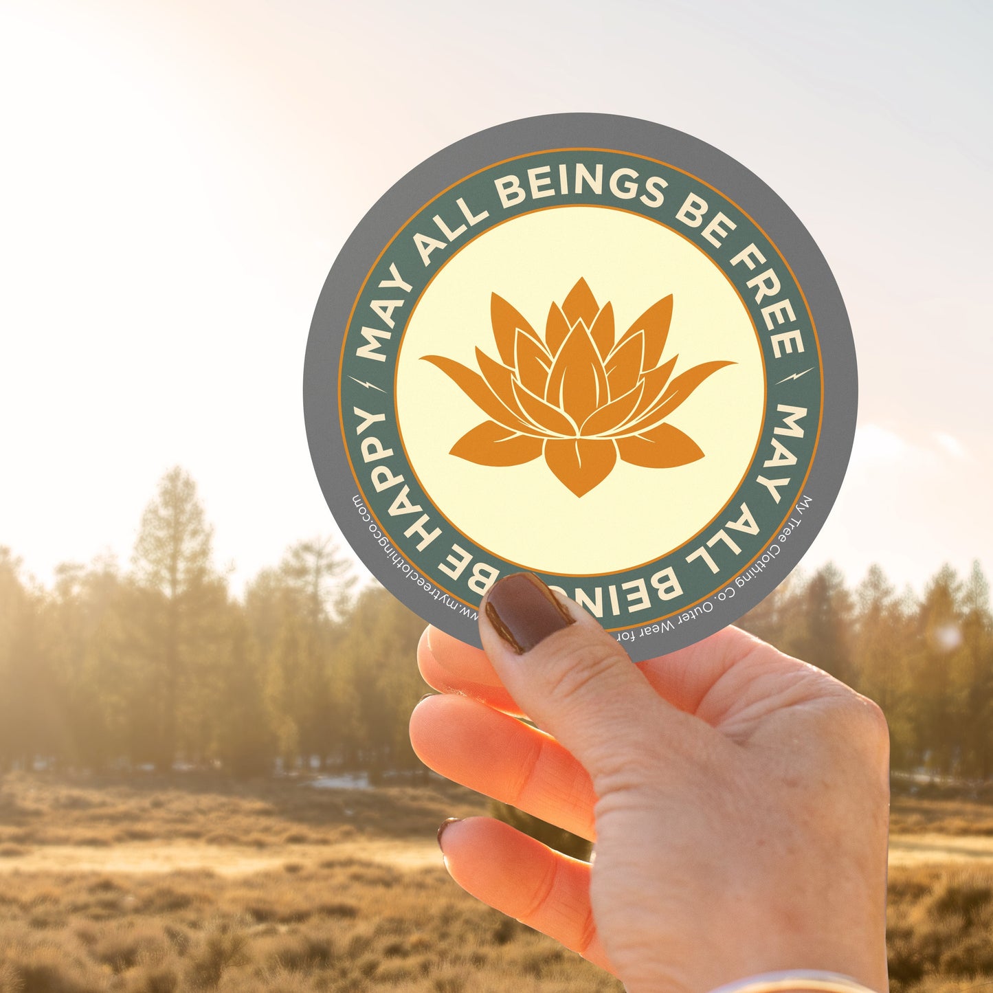 Hand holding a 4" circular sticker with a saffron colored lotus flower design and Mettā prayer, against a blurred natural background.