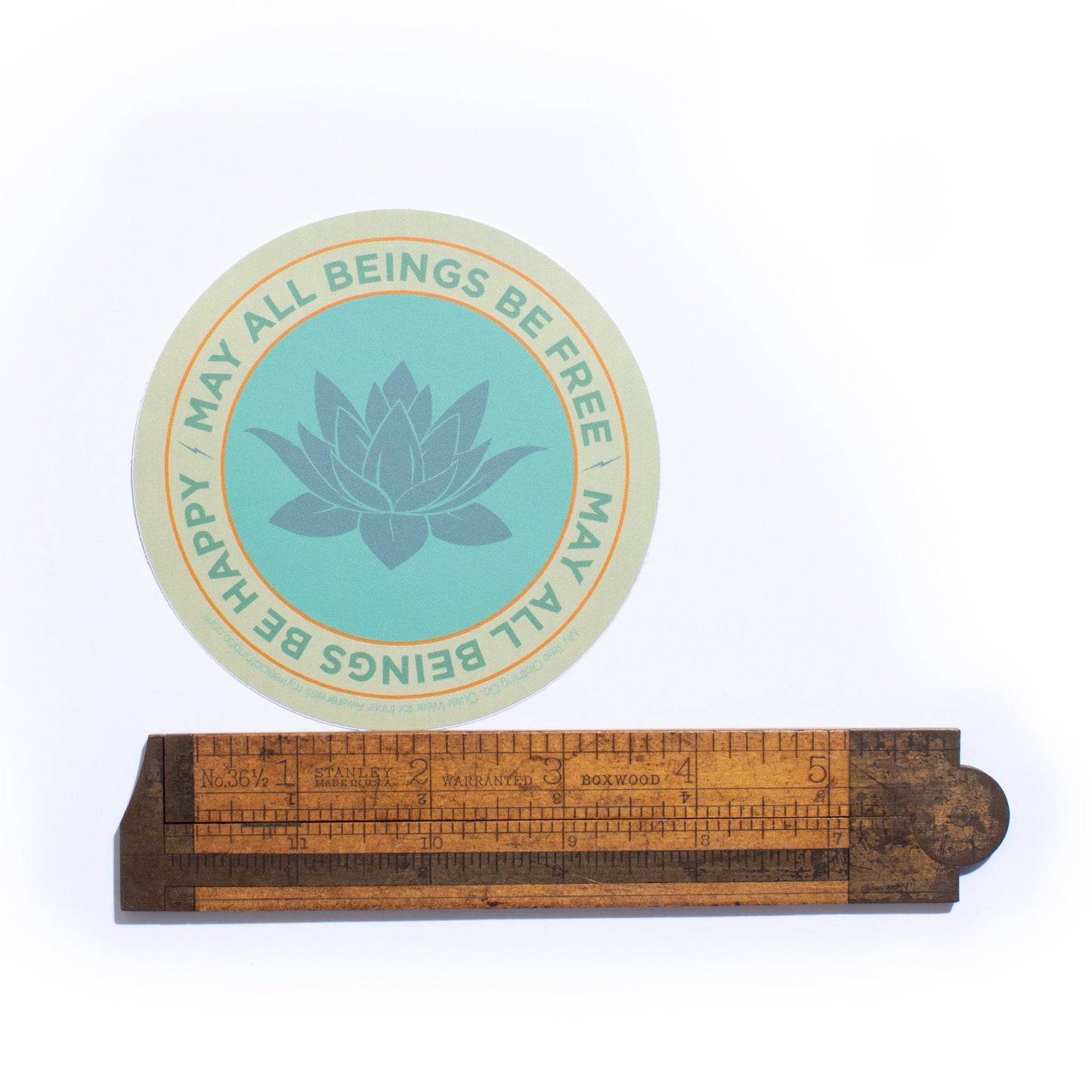 Round teal sticker with lotus flower design and text, may all beings be free may all beings be happy, on a wooden ruler stand.