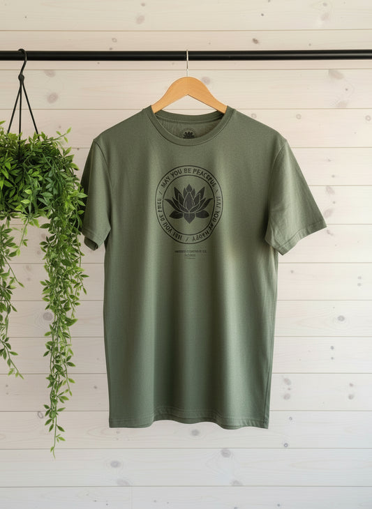 Green t-shirt with a black lotus and the metta prayer, may you be peaceful-may you be happy hanging on a black rod in front of a wooden wall.