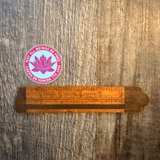 Wooden ruler on a wooden surface with a pink and purple sticker featuring a lotus flower design and the Buddhist mantra, may all beings be free-may all beings be happy.