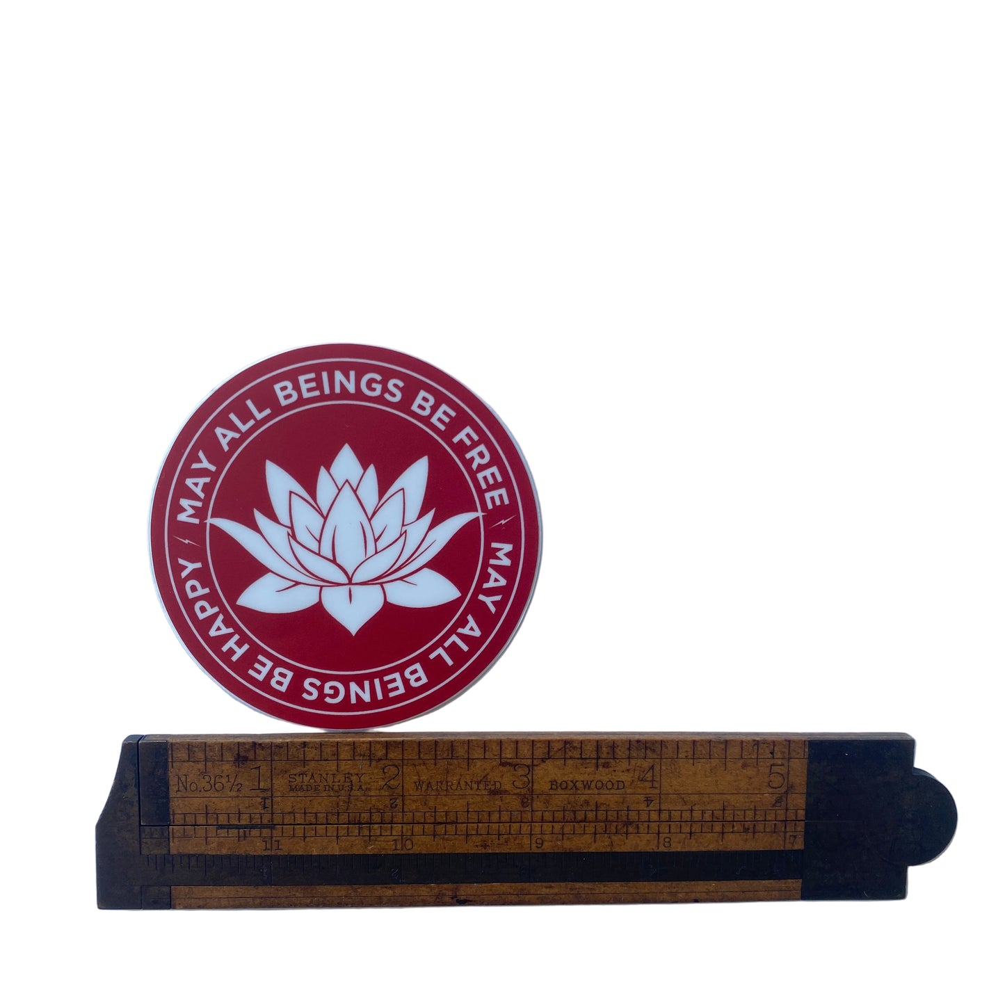 Round red and white three inch sticker with a lotus in the center and the Buddhist prayer, may all beings be happy-may all beings be free. It is being measured by an antique wooden ruler.