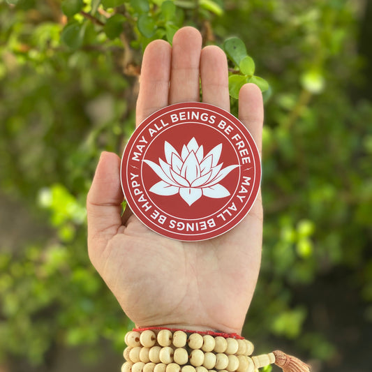 Round red and white three inch sticker being held in a hand with a lotus in the center and the Buddhist prayer, may all beings be happy-may all beings be free.