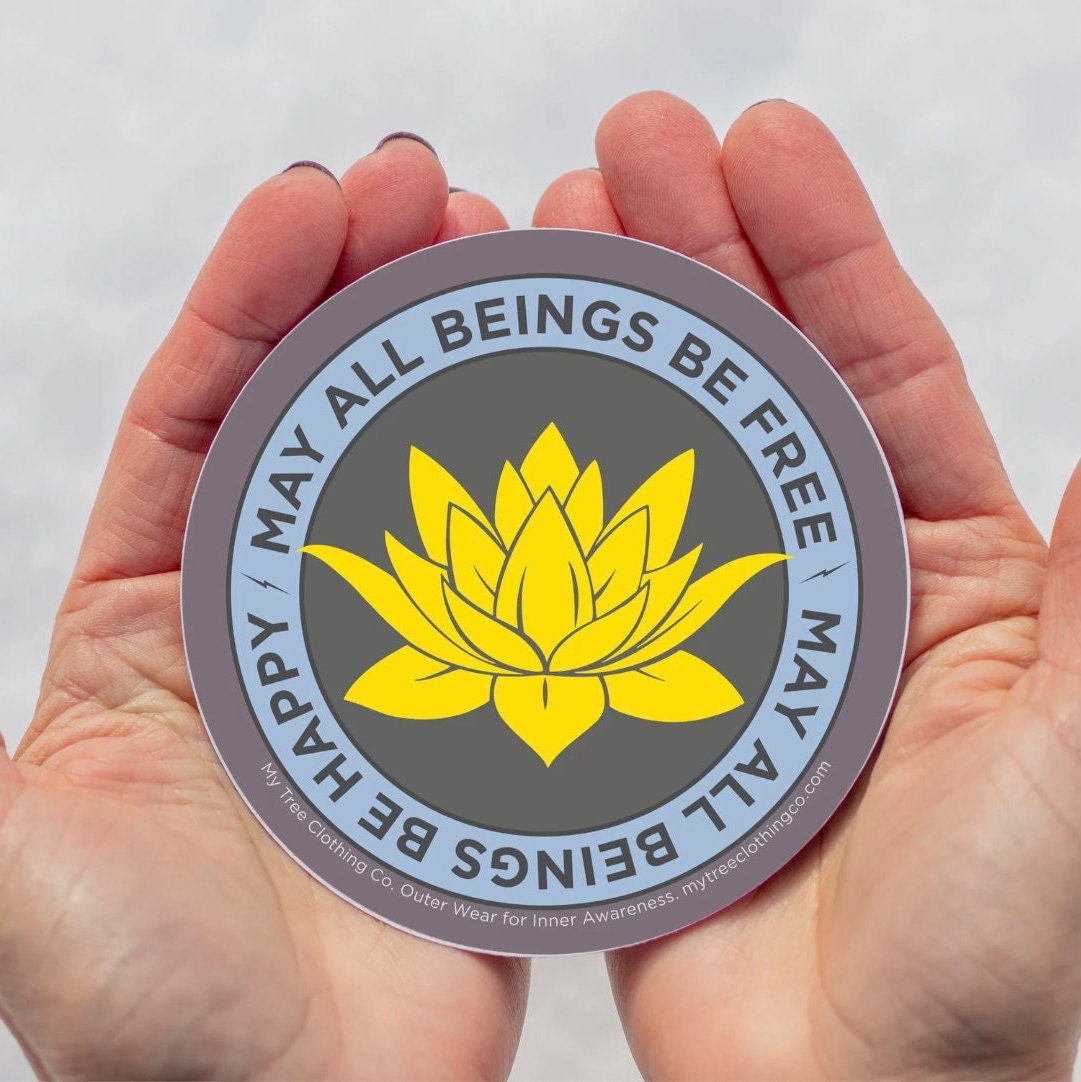 4" circular sticker with yellow lotus flower and text, 'may all beings be free may all beings be happy' held between two hands against a white background.