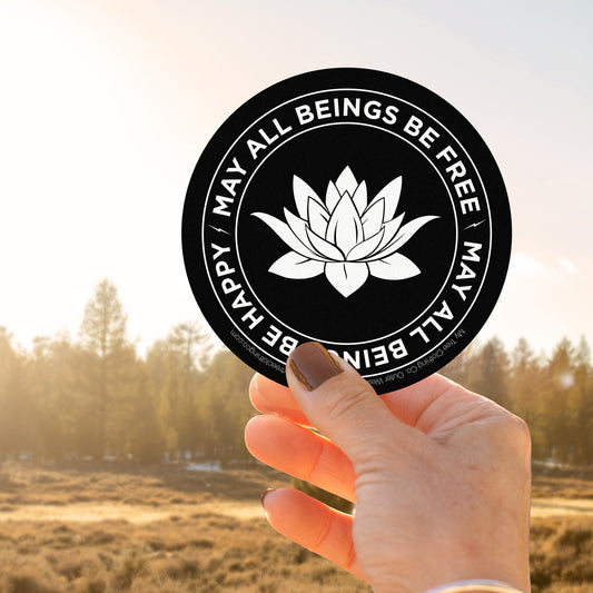 Hand holding a 4" black and white circular sticker with a lotus flower design and text, MAY ALL BEINGS BE FREE MAY ALL BEINGS BE HAPPY, against a scenic background.
