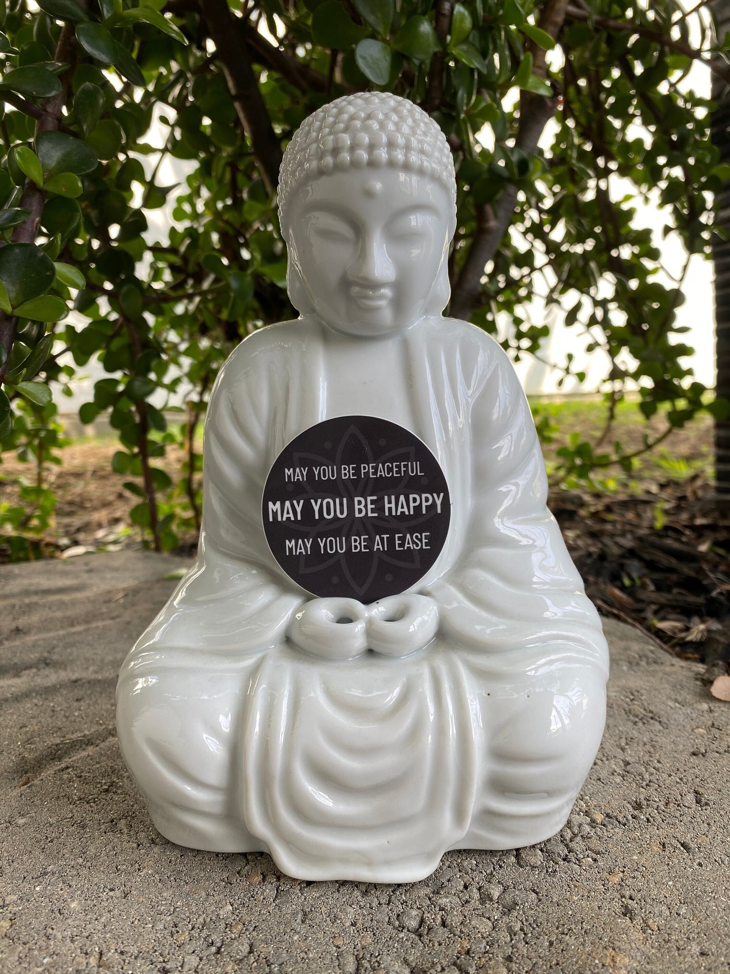 White Buddha statue with a 2" circular sticker with text, may you be peaceful may you be happy may you be at ease, outdoors.