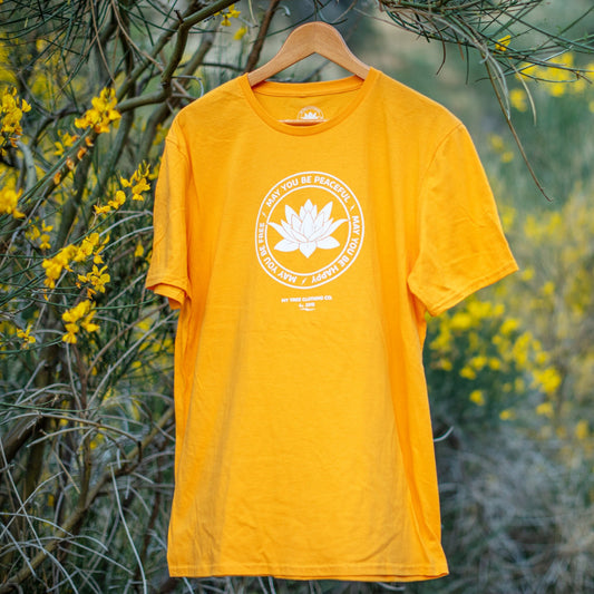 Yellow t-shirt with a white lotus flower graphic, may you be peaceful may you be happy may you be free, on a hanger against a natural background with yellow flowers.