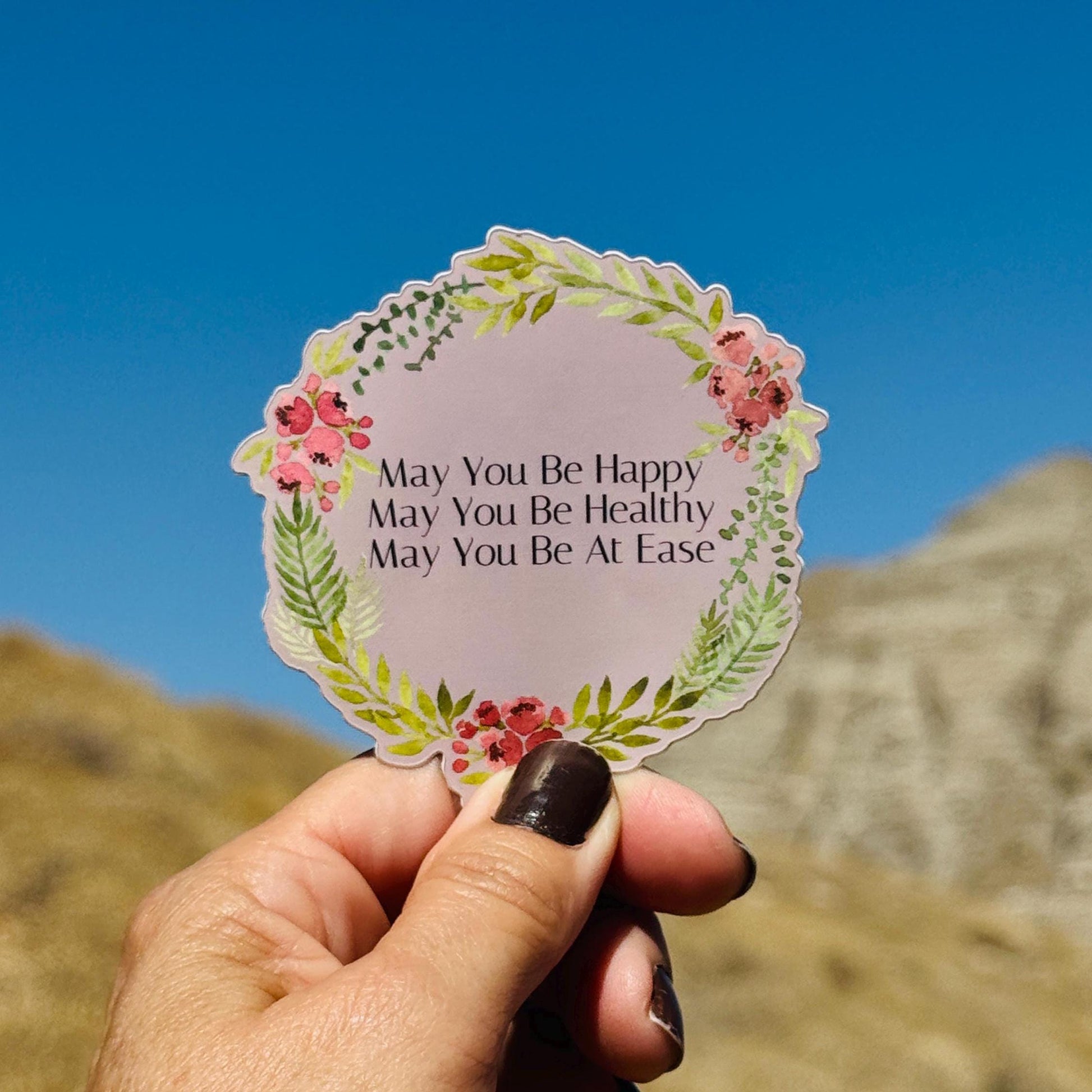 Hand holding a decorative sticker with text, may you be happy, may you be healthy, may you be at ease, against a mountainous background