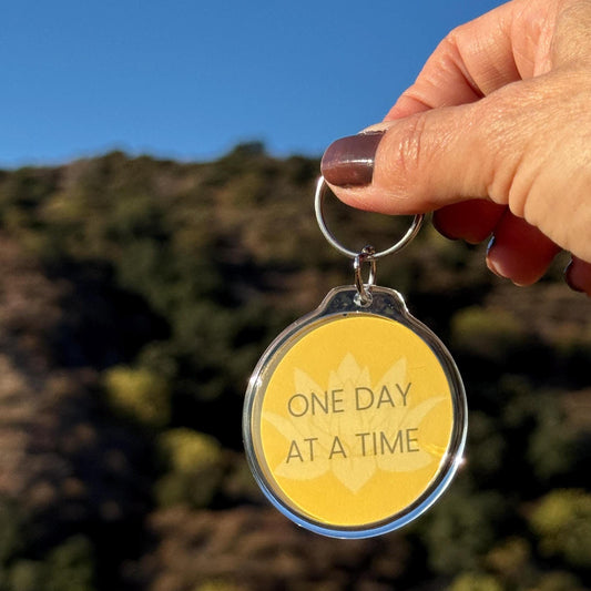 Hand holding a 2" yellow lotus keychain with 'One Day at a Time' text against a blurred natural background.