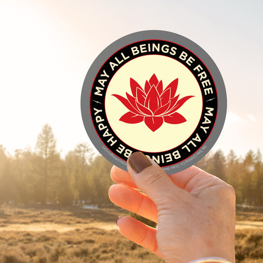 Hand holding a circular sticker with a red lotus flower design and text may all beings be free may all beings be happy, against a blurred natural background.
