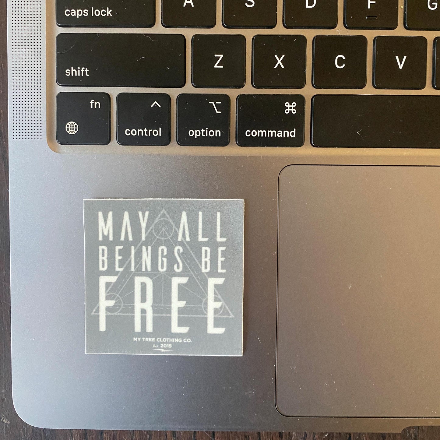 Laptop with a 2" square grey sticker with 'May all beings be free' text with sacred geometry design.