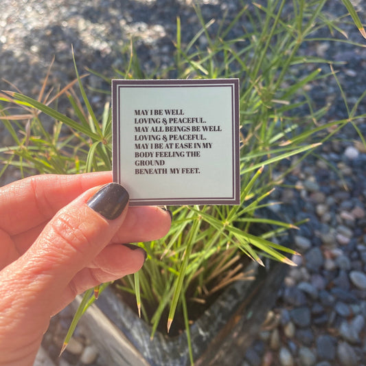 Hand holding a small sticker with text, may I be well, loving, and peaceful. "May all beings be well, loving, and peaceful. May I be at ease in my body feeling the ground beneath my feet" in front of a plant and pebbles.