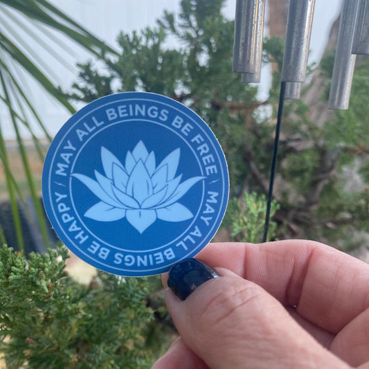 Blue sticker with lotus flower design and the text, 'may all beings be free may all beings be happy,' held by a hand against a blurred green background.