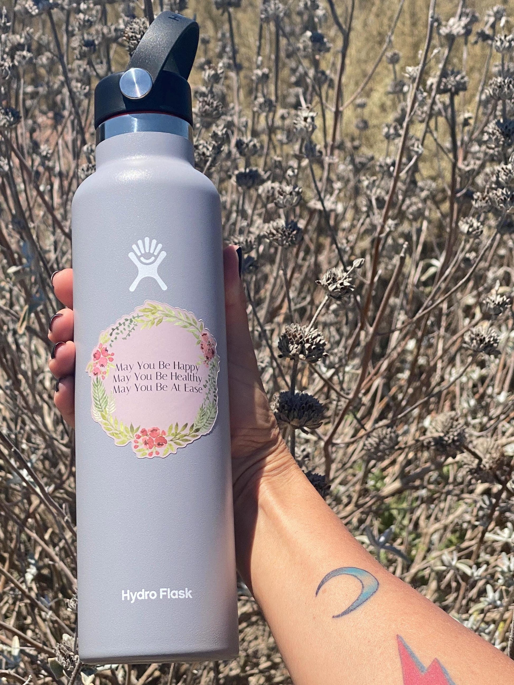 Hydro Flask water bottle with a floral sticker that reads, may you be happy, may you be healthy, may you be at ease, held in front of dry plants.