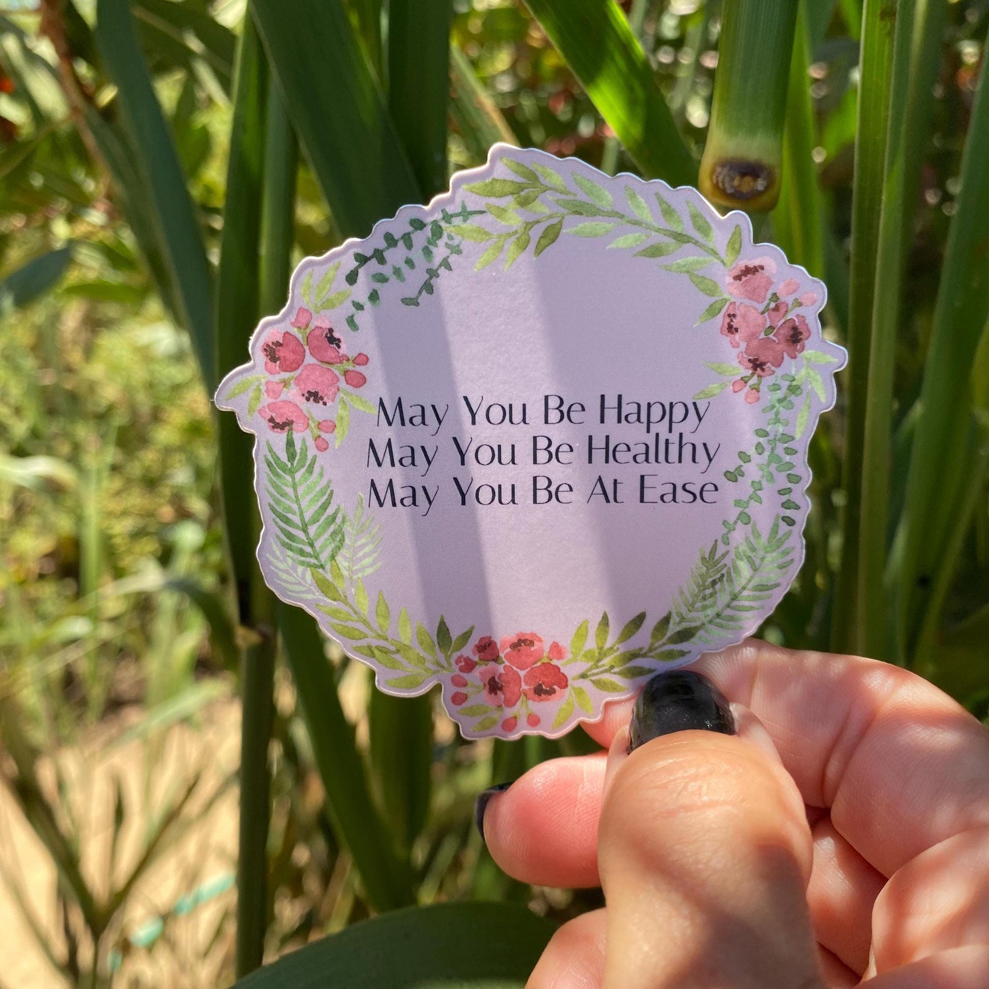 Decorative sticker with floral design and text, may you be happy, may you be healthy, may you be at ease, held in front of green plants