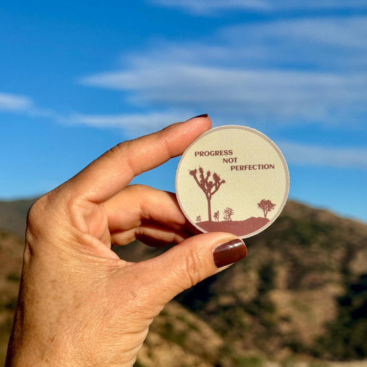 Hand holding a circular sticker with desert landscape design and 'Progress Not Perfection' text against a mountainous background.