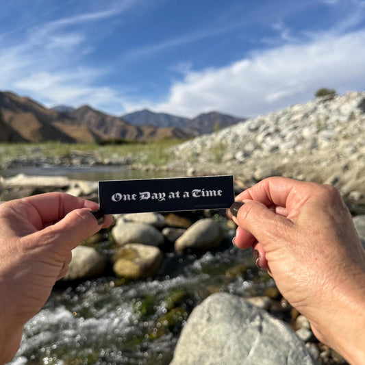 Person holding a black and white, old english font 'One Day at a Time' sticker, with mountains in the background.