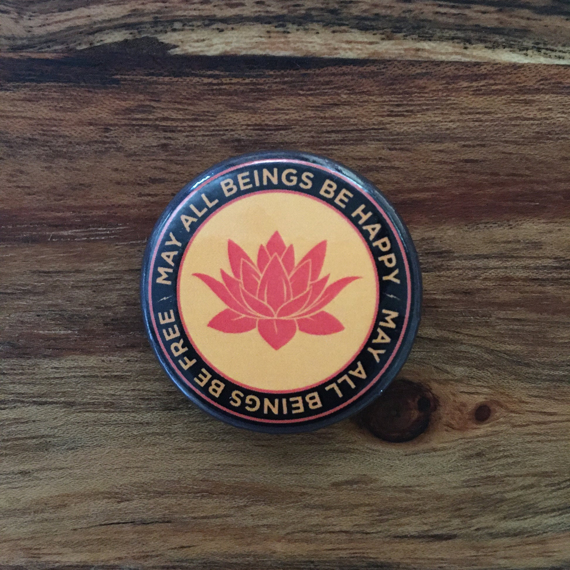 1.25" Orange round pin back button with a buddhist lotus flower and the text 'may all beings be happy may all beings be free,' on a wooden surface.