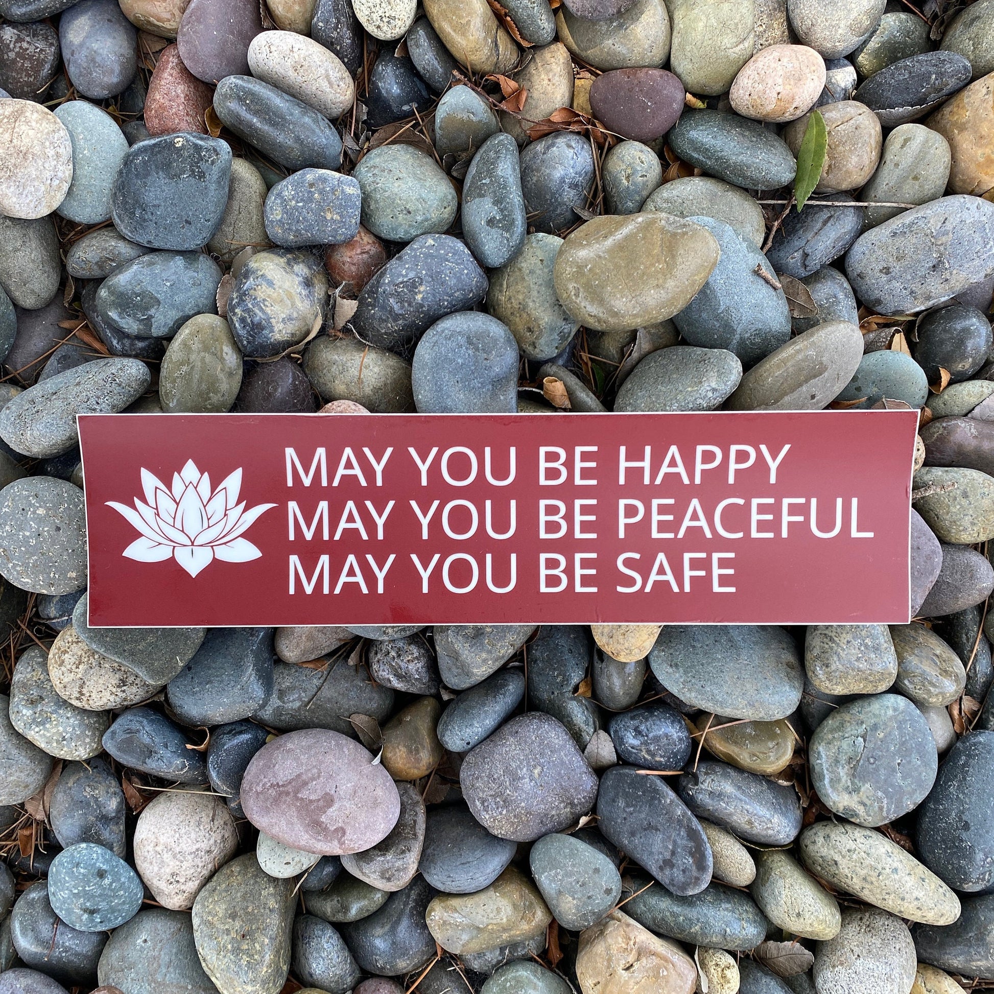 Red and white bumper sticker with a lotus and the prayer, may you be happy may you be peaceful may you be safe, on a bed of multicolored stones.