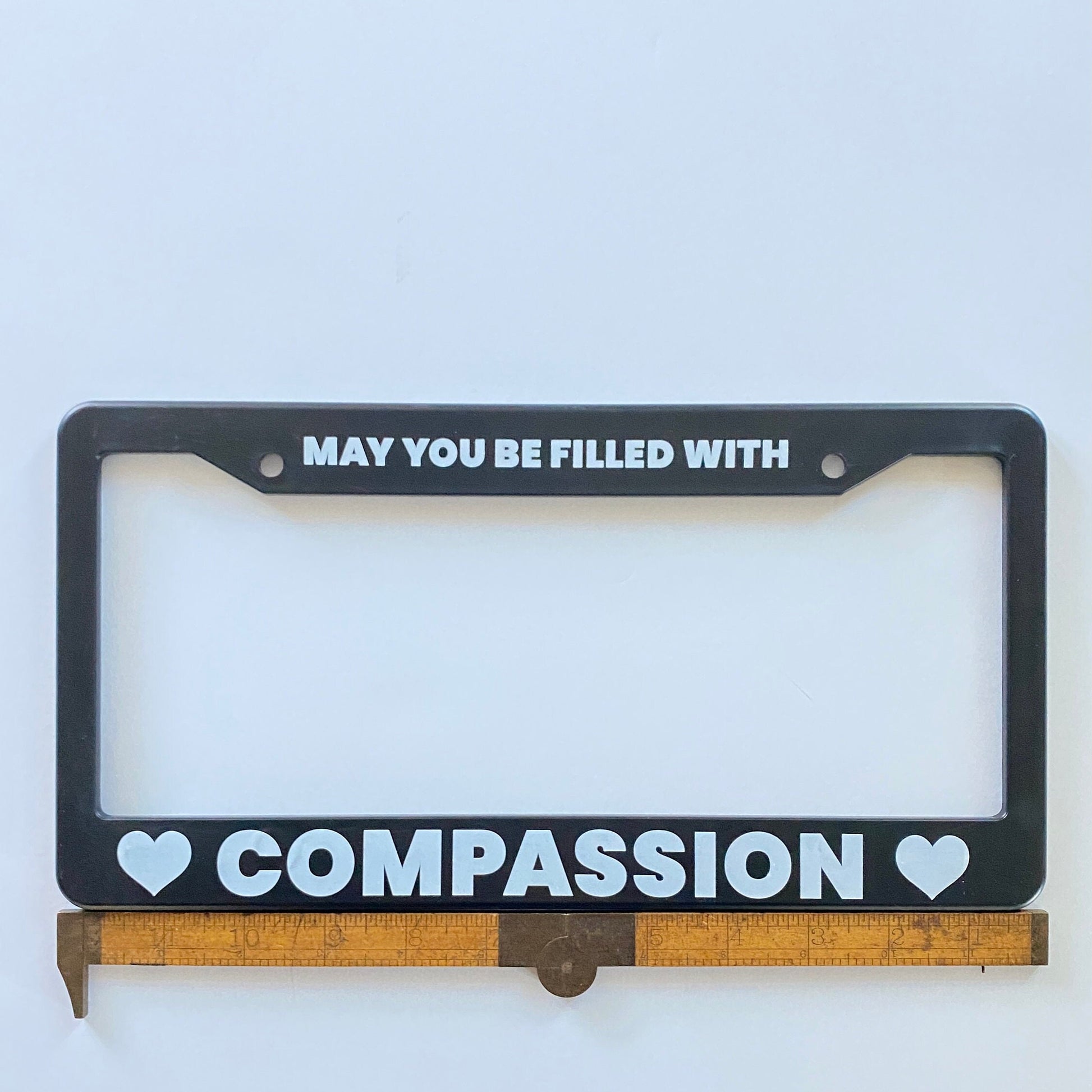 Black license plate frame with text 'MAY YOU BE FILLED WITH COMPASSION' on a white background, being measured by an antique ruler.