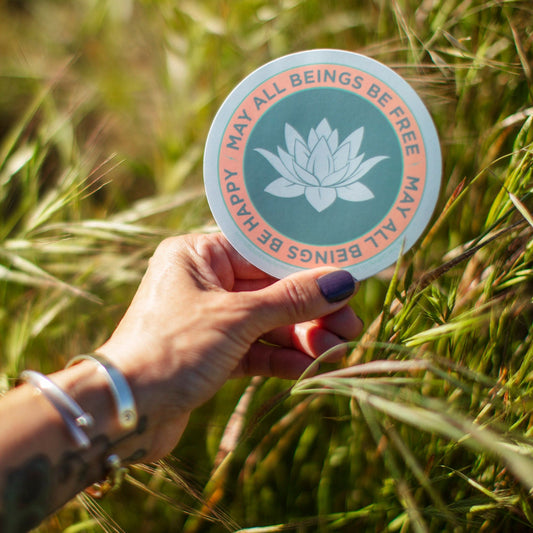 4" circular sticker with a lotus flower design and text, may all beings be free may all beings be happy in a grassy field.