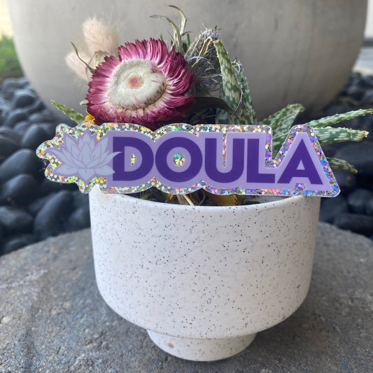 White pot with flowers and a purple 'DOULA' sticker with glittery effect on a stone surface.
