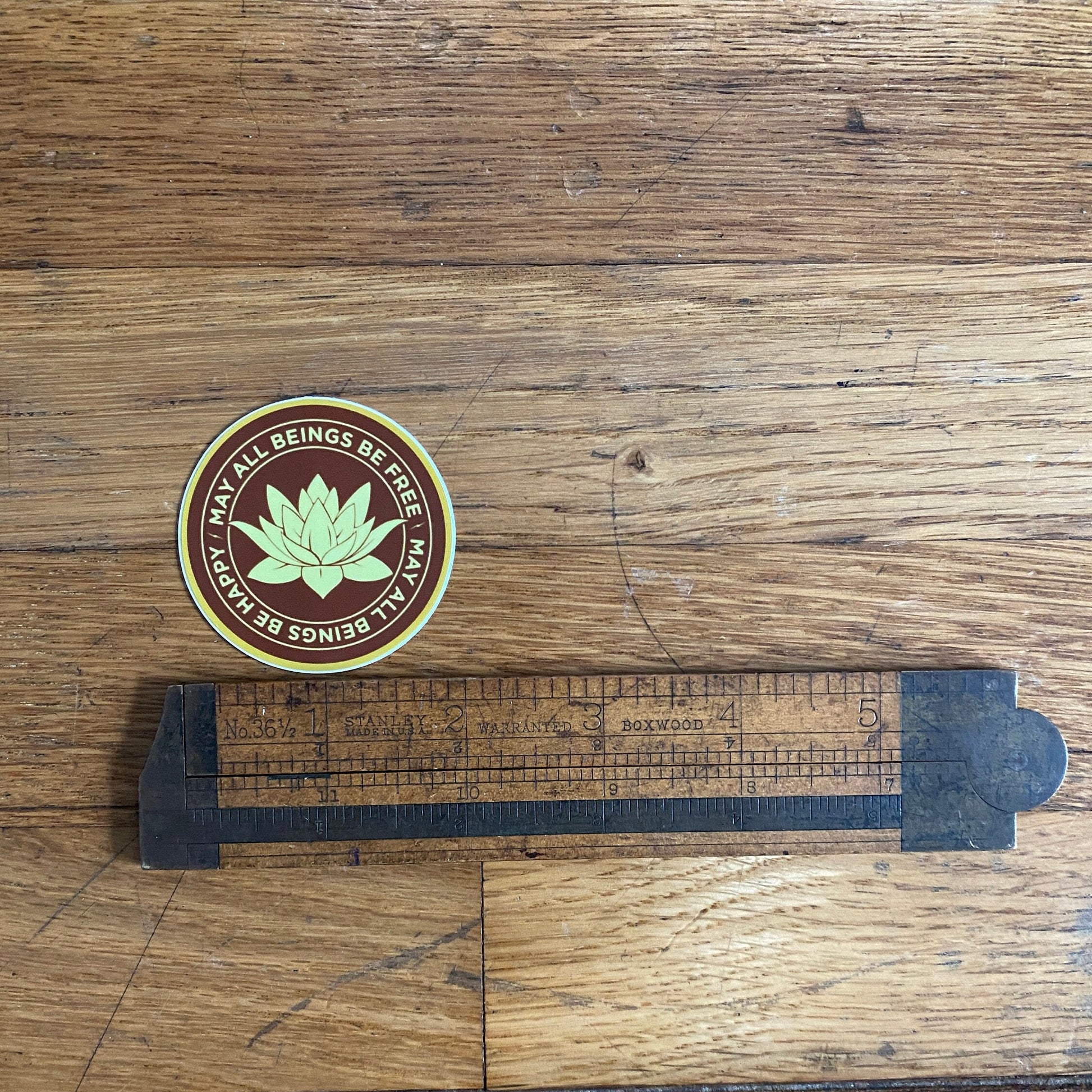 2" round saffron and gold vinyl sticker with a Buddhist mettā prayer text, may all beings be free, may all being be happy, on a wooden surface with a ruler for scale.