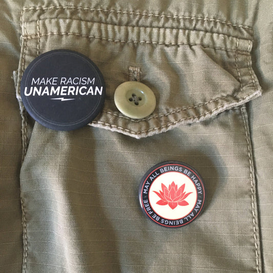 Two buttons on a beige jacket, one with 'Make Racism Unamerican' and one 1.25" pin back button with a red lotus flower design and the text, may all beings be free may all beings be happy.
