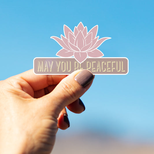 Hand holding a sticker with a lotus flower and 'May You Be Peaceful' text against a blue background