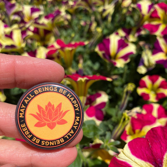 Orange round pin back button with a buddhist lotus flower and the text 'may all beings be happy may all beings be free', design held in front of colorful flowers.