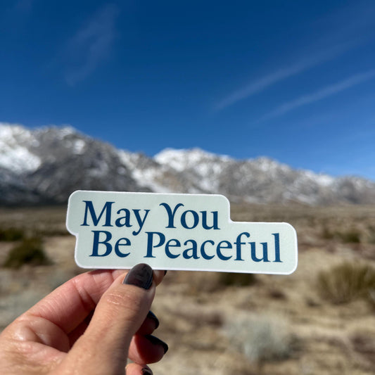 Hand holding a light blue and dark blue 'May You Be Peaceful' sticker with mountains in the background.