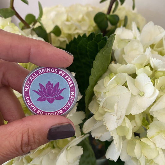 Hand holding a 1.25" round pin back button with a purple lotus design and text, may all beings be free may all beings be happy.